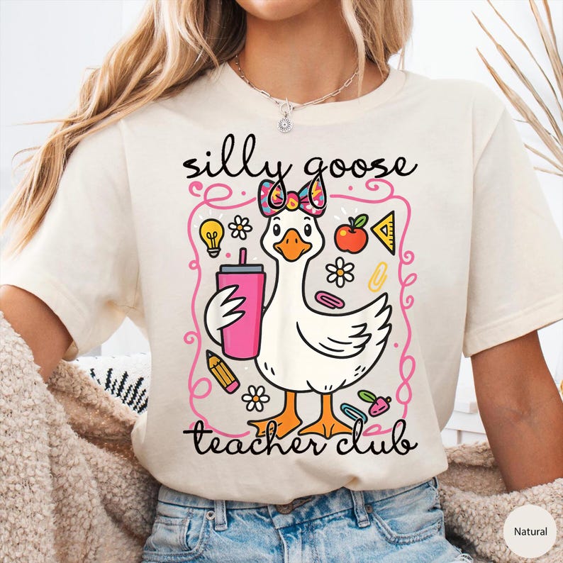 Puede incluir: Camiseta color crema con un dise&ntilde;o de ganso de dibujos animados con un lazo y sosteniendo una bebida rosa. El texto dice "silly goose teacher club". El dise&ntilde;o incluye elementos escolares como una bombilla, una manzana y un l&aacute;piz.