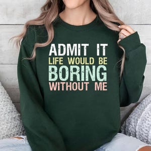 May include: Dark green sweatshirt with the text "ADMIT IT LIFE WOULD BE BORING WITHOUT ME" in white, yellow, light blue, and pink. The sweatshirt is a long-sleeved crew neck.