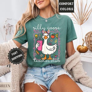 Puede incluir: Camiseta verde claro con un dibujo de un ganso sosteniendo una bebida rosa, rodeado de ilustraciones escolares. El texto "silly goose teacher club" se muestra debajo. La camiseta es de la colecci&oacute;n comfort colors.
