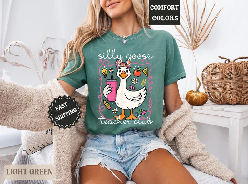Puede incluir: Camiseta verde claro con un dibujo de un ganso sosteniendo una bebida rosa, rodeado de ilustraciones escolares. El texto "silly goose teacher club" se muestra debajo. La camiseta es de la colecci&oacute;n comfort colors.