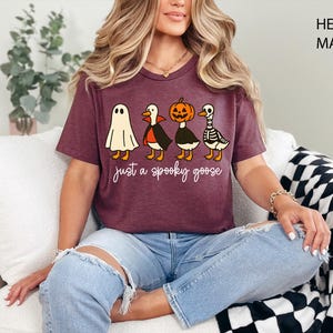 以下が含まれることがあります： ハロウィーンをテーマにしたグラフィックが特徴の、ヘザーマルーンのTシャツ。デザインは、お化け、吸血鬼、カボチャ、スケルトンに扮した4羽の漫画のガチョウが特徴です。「just a spooky goose」というテキストがガチョウの下にあります。