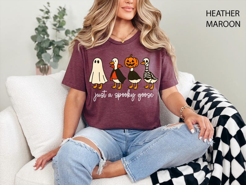 以下が含まれることがあります： ハロウィーンをテーマにしたグラフィックが特徴の、ヘザーマルーンのTシャツ。デザインは、お化け、吸血鬼、カボチャ、スケルトンに扮した4羽の漫画のガチョウが特徴です。「just a spooky goose」というテキストがガチョウの下にあります。