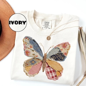 May include: Ivory-colored sweatshirt featuring a patchwork butterfly design. The butterfly wings are made of various patterned fabrics in shades of blue, pink, yellow, and red. A gold necklace with a pendant is draped on the shirt.