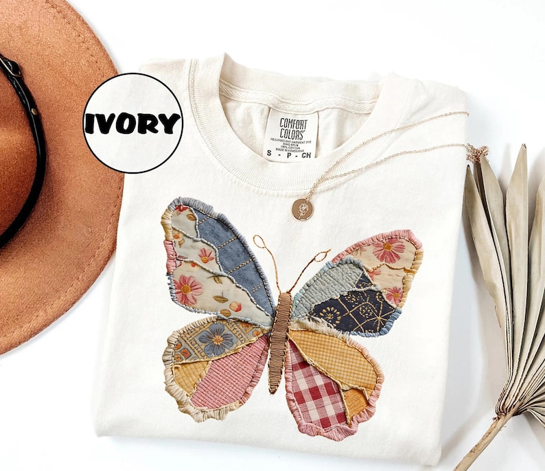 May include: Ivory-colored sweatshirt featuring a patchwork butterfly design. The butterfly wings are made of various patterned fabrics in shades of blue, pink, yellow, and red. A gold necklace with a pendant is draped on the shirt.