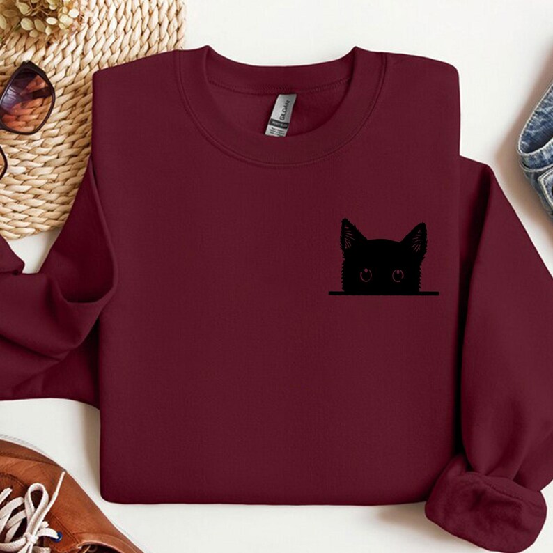 May include: A maroon long-sleeve sweatshirt featuring a black graphic of a cat head peeking over a horizontal line on the left chest area. The garment has a crew neckline and a standard fit, showcasing a simple design on a solid dark red fabric.