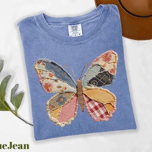 May include: A blue t-shirt with a patchwork butterfly design. The butterfly wings are made of various patterned fabrics in shades of blue, pink, yellow, and red. The shirt has a crew neck and short sleeves.