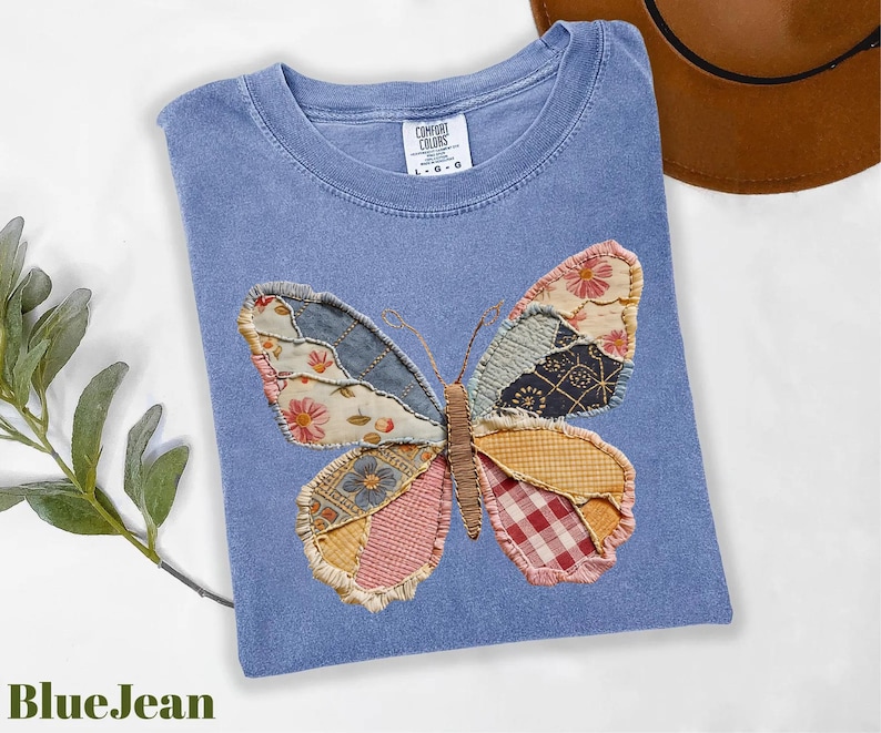 May include: A blue t-shirt with a patchwork butterfly design. The butterfly wings are made of various patterned fabrics in shades of blue, pink, yellow, and red. The shirt has a crew neck and short sleeves.
