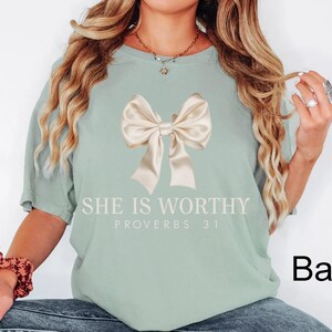 May include: A light green t-shirt with a large beige bow and the text "She is Worthy Proverbs 31" printed on the front.