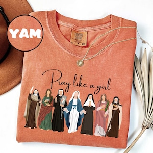 May include: A coral-colored t-shirt with the phrase "Pray like a girl" and an illustration of several women in religious attire. A gold necklace and a brown hat are also visible. The shirt has a Comfort Colors tag and the word "YAM" in a circle.