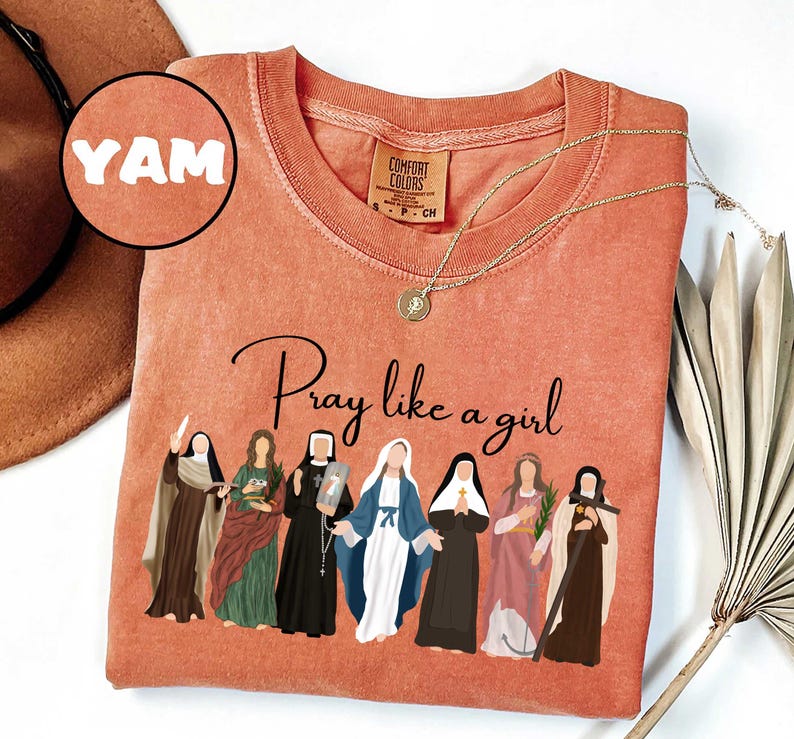 May include: A coral-colored t-shirt with the phrase "Pray like a girl" and an illustration of several women in religious attire. A gold necklace and a brown hat are also visible. The shirt has a Comfort Colors tag and the word "YAM" in a circle.