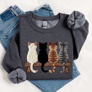 May include: A dark grey sweatshirt with an embroidered design of four cats sitting on a wooden bench. The cats are white, black, brown, and grey. The sweatshirt is displayed with blue denim jeans.