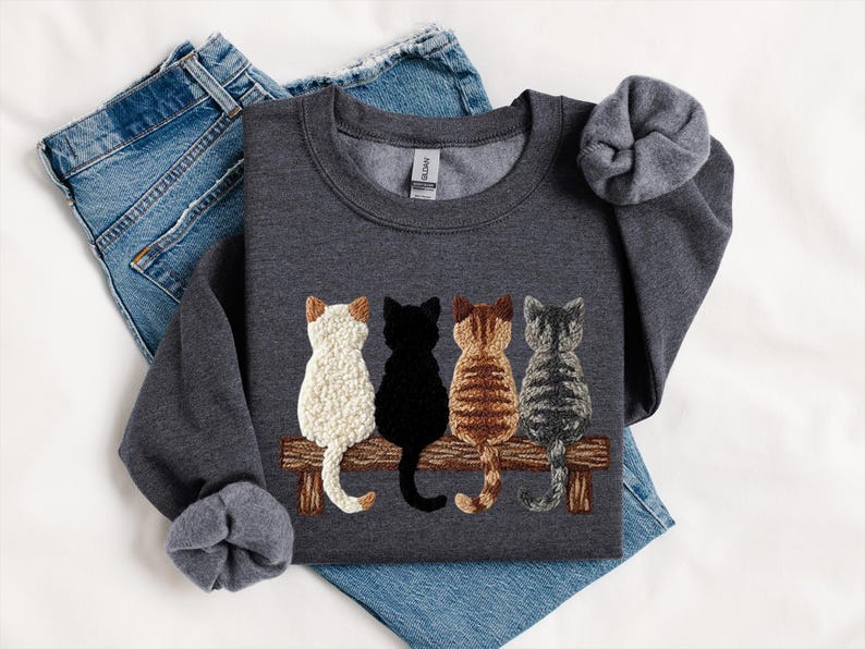 May include: A dark grey sweatshirt with an embroidered design of four cats sitting on a wooden bench. The cats are white, black, brown, and grey. The sweatshirt is displayed with blue denim jeans.