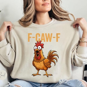 May include: A sand-colored crewneck sweatshirt featuring a graphic of a surprised brown rooster with a red comb and wattle. Above the rooster, the text F-CAW-F is printed in a bold, yellow, distressed font. The sweatshirt has long sleeves.