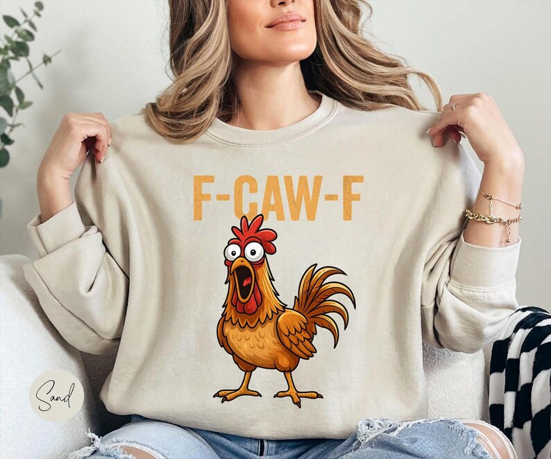 May include: A sand-colored crewneck sweatshirt featuring a graphic of a surprised brown rooster with a red comb and wattle. Above the rooster, the text F-CAW-F is printed in a bold, yellow, distressed font. The sweatshirt has long sleeves.