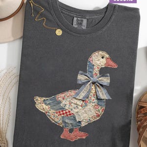 Peut inclure: Un t-shirt gris fonc&eacute; Comfort Colors avec un motif d'oie en patchwork et un n&oelig;ud en vichy bleu. L'oie est faite de divers tissus &agrave; motifs rouges, bleus et cr&egrave;me. Un collier en or avec un pendentif est drap&eacute; sur le t-shirt.