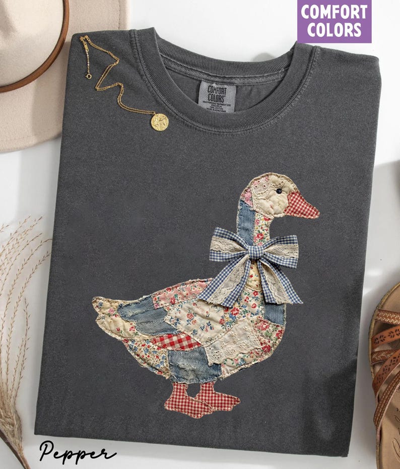 Peut inclure: Un t-shirt gris fonc&eacute; Comfort Colors avec un motif d'oie en patchwork et un n&oelig;ud en vichy bleu. L'oie est faite de divers tissus &agrave; motifs rouges, bleus et cr&egrave;me. Un collier en or avec un pendentif est drap&eacute; sur le t-shirt.