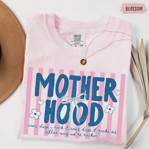 May include: A light pink t-shirt with the word "MOTHERHOOD" in blue, decorated with a bow, hearts, and flowers. The shirt has pink vertical stripes and the text "some days i rock it, some days it rocks me either way we're rockin'".