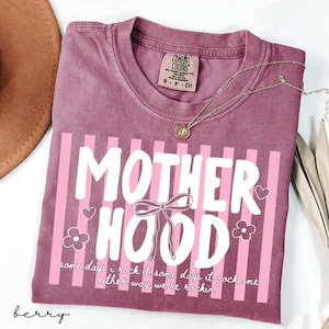 May include: Berry-colored t-shirt with the word "MOTHERHOOD" in white, accented with pink stripes, a bow, hearts, and flowers. The text "some days I rock it, some days it rocks me, either way we're rockin" is also printed on the shirt. A gold necklace is draped across the shirt.
