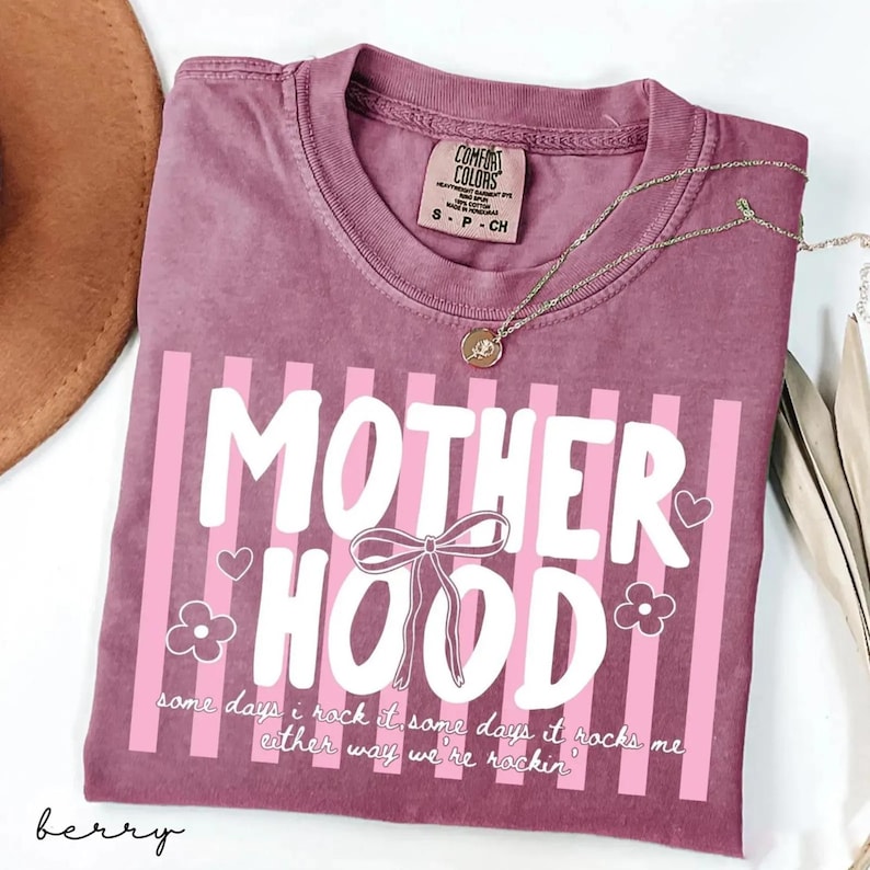 May include: Berry-colored t-shirt with the word "MOTHERHOOD" in white, accented with pink stripes, a bow, hearts, and flowers. The text "some days I rock it, some days it rocks me, either way we're rockin" is also printed on the shirt. A gold necklace is draped across the shirt.
