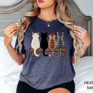 May include: Heather navy t-shirt with an embroidered design of four cats on a wooden bench. The cats are white, black, brown, and grey. The words "HEATHER NAVY" are printed on the bottom right of the image.
