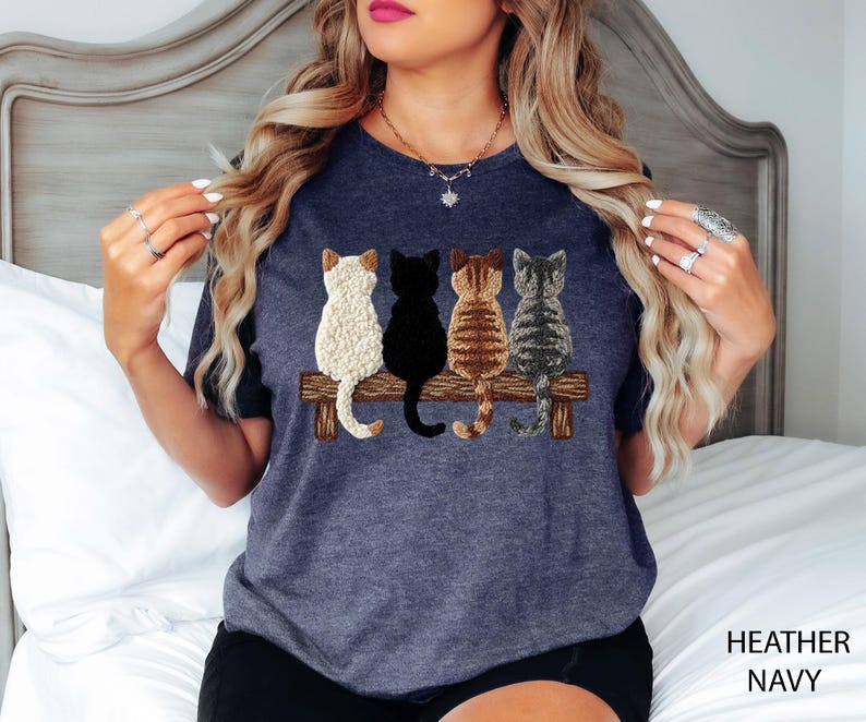 May include: Heather navy t-shirt with an embroidered design of four cats on a wooden bench. The cats are white, black, brown, and grey. The words "HEATHER NAVY" are printed on the bottom right of the image.