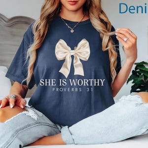 May include: A navy blue t-shirt with a white bow and the text "She is Worthy Proverbs 31" printed on the front. The model is wearing the shirt with denim jeans.