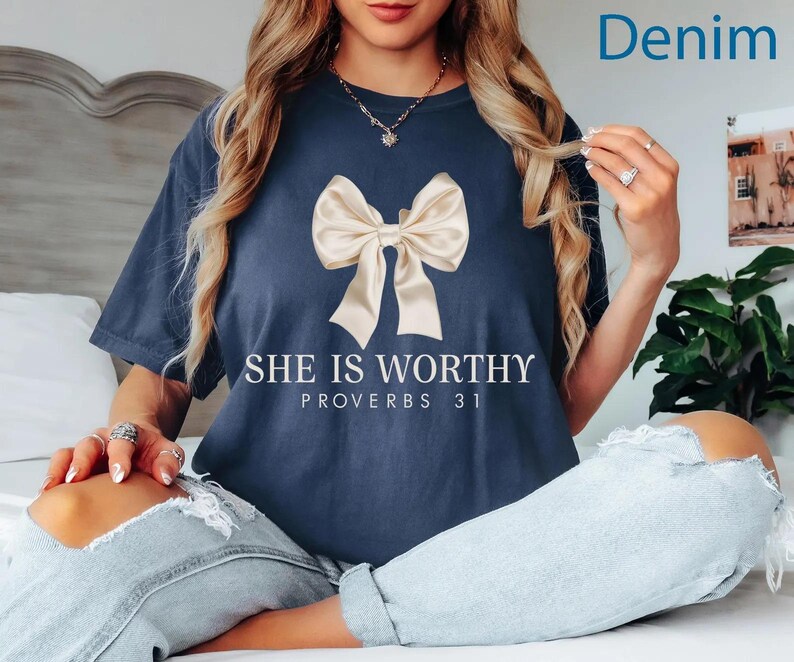 May include: A navy blue t-shirt with a white bow and the text "She is Worthy Proverbs 31" printed on the front. The model is wearing the shirt with denim jeans.