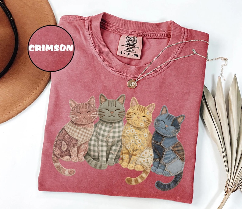 May include: Crimson-colored t-shirt featuring four smiling cat illustrations with patchwork designs. The cats are in various colors and patterns, including paisley, gingham, and floral. A silver necklace with a pendant is draped on the shirt.