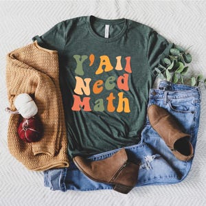 K&ouml;nnte beinhalten: Ein gr&uuml;nes T-Shirt mit dem Text "Y'all Need Math" in bunten, Retro-Lettern.
