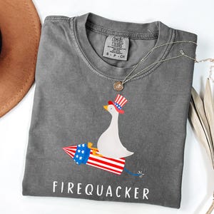 K&ouml;nnte beinhalten: Ein anthrazitfarbenes T-Shirt mit einer Grafik einer wei&szlig;en Gans, die einen rot-wei&szlig;-blauen Zylinder tr&auml;gt und auf einer Feuerwerksrakete reitet. Der Text "FIREQUACKER" ist in Wei&szlig; unter der Grafik gedruckt. Mit Comfort Colors Etikett.