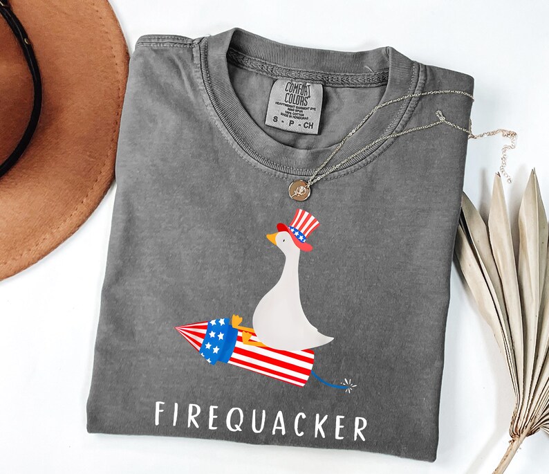 K&ouml;nnte beinhalten: Ein anthrazitfarbenes T-Shirt mit einer Grafik einer wei&szlig;en Gans, die einen rot-wei&szlig;-blauen Zylinder tr&auml;gt und auf einer Feuerwerksrakete reitet. Der Text "FIREQUACKER" ist in Wei&szlig; unter der Grafik gedruckt. Mit Comfort Colors Etikett.