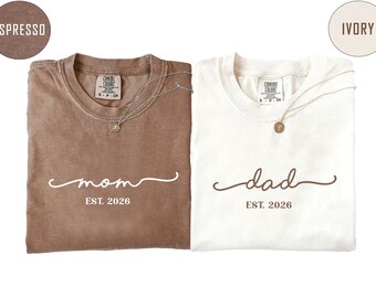 Puede incluir: Dos camisetas de manga corta con cuello redondo. La camiseta marrón espresso presenta el texto blanco "mom" en letra cursiva sobre "EST. 2026". La camiseta color marfil presenta el texto marrón "dad" en letra cursiva sobre "EST. 2026".