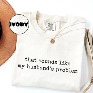 May include: Ivory Comfort Colors t-shirt with the text "that sounds like my husband's problem" in black. A gold necklace with a pendant rests on the shirt. A brown hat and dried plant are in the background.