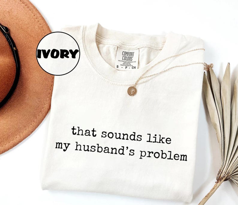 May include: Ivory Comfort Colors t-shirt with the text "that sounds like my husband's problem" in black. A gold necklace with a pendant rests on the shirt. A brown hat and dried plant are in the background.