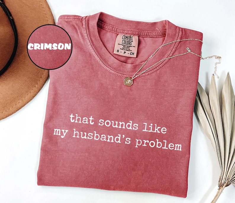 May include: Crimson t-shirt with the white text "that sounds like my husband's problem". A silver necklace with a pendant is on the shirt. The shirt is folded on a white surface. The shirt is a Comfort Colors brand.