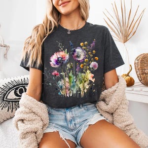 May include: Dark gray t-shirt featuring a watercolor floral design with purple, pink, and yellow flowers. The shirt has a crew neck and short sleeves. The model is wearing denim shorts and a cream-colored jacket.