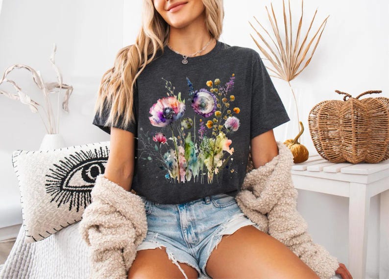 May include: Dark gray t-shirt featuring a watercolor floral design with purple, pink, and yellow flowers. The shirt has a crew neck and short sleeves. The model is wearing denim shorts and a cream-colored jacket.