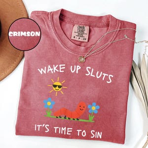 Puede incluir: Camiseta Comfort Colors carmes&iacute; con el texto "WAKE UP SLUTS IT'S TIME TO SIN" y un gr&aacute;fico de un sol sonriente, un gusano y flores. Un collar dorado est&aacute; drapeado sobre la camisa.