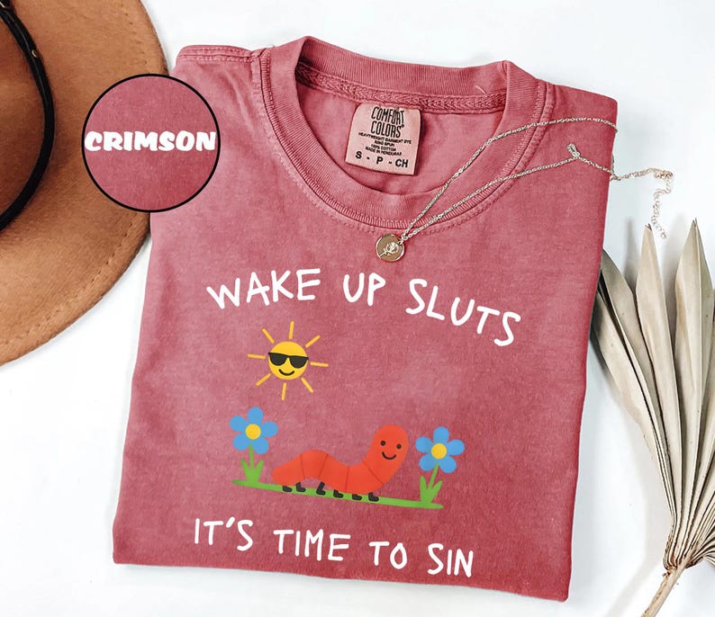 Puede incluir: Camiseta Comfort Colors carmes&iacute; con el texto "WAKE UP SLUTS IT'S TIME TO SIN" y un gr&aacute;fico de un sol sonriente, un gusano y flores. Un collar dorado est&aacute; drapeado sobre la camisa.