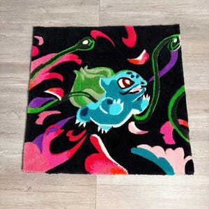 Könnte beinhalten: Ein quadratischer, schwarzer Teppich mit einer Cartoon-Figur von Bulbasaur in Blau- und Grüntönen. Der Teppich hat ein farbenfrohes Design mit rosa, roten und lila Akzenten. Der Teppich ist ein dekorativer Artikel.