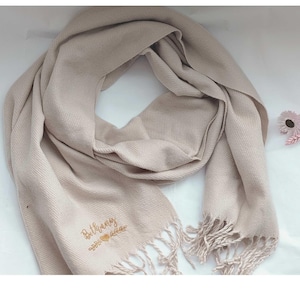 May include: A beige scarf with fringe detailing. The scarf is embroidered with the name "Bethany" in a cursive font, accompanied by a heart and leaf design. The scarf is made of a soft material and is draped.