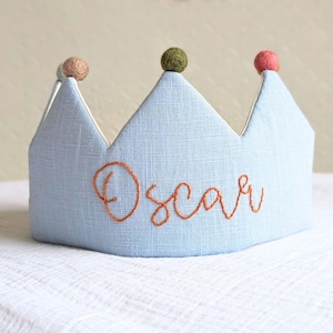 Può includere: Una corona di stoffa azzurra con il nome "Oscar" ricamato in filo arancione. La corona ha tre punte, ognuna sormontata da un piccolo pompon colorato. Corona per bambini.