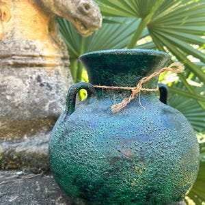 May include: A textured, dark green ceramic vase with two handles and a rope tied around the neck. The vase has a rounded body and a slightly flared rim. The background includes a stone statue and green foliage.