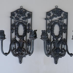 May include: Pair of ornate, dark gray metal wall sconces. Each sconce features a floral design, a central wreath, and two candle holders. The sconces are mounted on a white wall, showcasing their intricate details and vintage style.