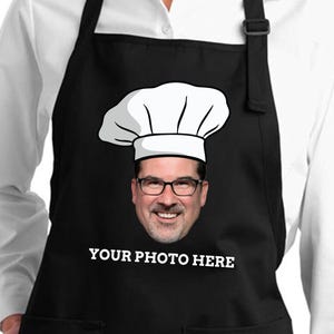 May include: Black apron with a chef hat graphic and a smiling face wearing glasses. The text "YOUR PHOTO HERE" is printed in white at the bottom. The apron is worn over a white collared shirt.