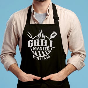 May include: Black apron with white text that reads "Grill Master William" and a grill design. The apron has two white grilling tools on either side of the design.