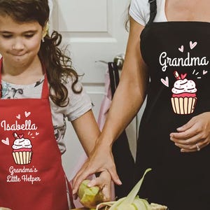 May include: A red apron with the name "Isabella" and the text "Grandma's Little Helper" printed on it. The apron features a cupcake graphic with hearts. A black apron with the word "Grandma" and a cupcake graphic is also visible.
