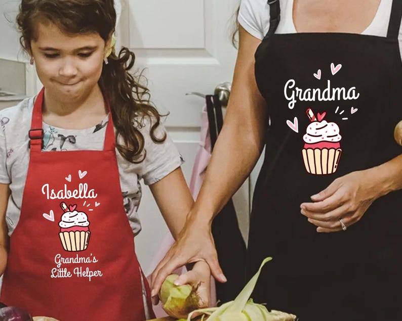 May include: A red apron with the name "Isabella" and the text "Grandma's Little Helper" printed on it. The apron features a cupcake graphic with hearts. A black apron with the word "Grandma" and a cupcake graphic is also visible.
