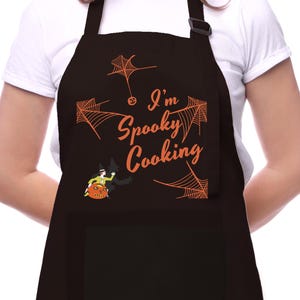 May include: Black apron with an orange spiderweb design and the text "I'm Spooky Cooking". A cartoon witch and pumpkin are also featured on the apron.