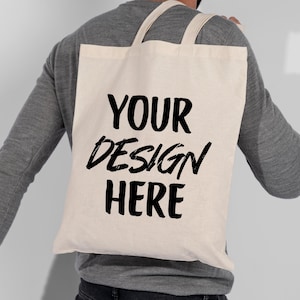 K&ouml;nnte beinhalten: Eine cremefarbene Tragetasche mit den Worten "YOUR DESIGN HERE" in fetter schwarzer Schrift. Die Tasche hat zwei Tr&auml;ger und wird &uuml;ber der Schulter getragen. Die Tasche ist aus einem robusten Stoff gefertigt.